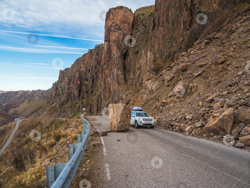 Скачать Белый внедорожник Rover объезжает большую скалу по узкой опасной горной дороге. Опасное вождение по горной дороге во время камнепада. фотосток Ozero