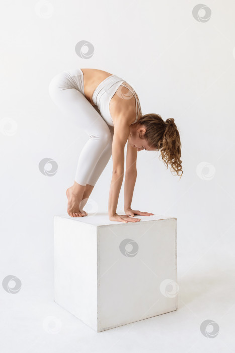 Скачать Молодая женщина, практикующая йогу, делает подготовительный выход к упражнению эка пада бхуджапидасана, готовится к стойке на руках, тренируется на белом кубе в спортивной одежде на светлом фоне фотосток Ozero