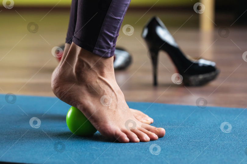Скачать Женщина выполняет массаж мышц стоп массажным шариком, стоя на коврике в студии. Профилактика усталости ног, вздутия вен после ношения обуви на каблуках фотосток Ozero