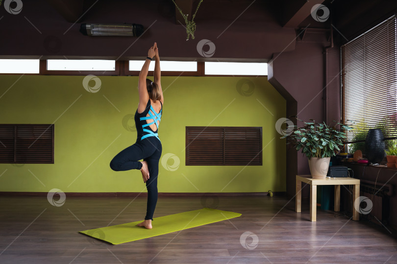 Скачать Женщина в спортивном комбинезоне, выполняет вариацию упражнения врикшасана, поза дерева, стоит с поднятыми руками на коврике в студии. фотосток Ozero
