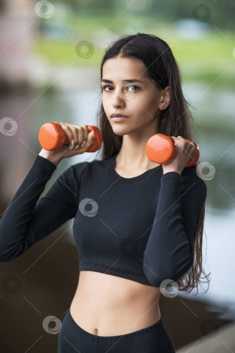 Скачать Портрет молодой красивой спортивной девушки, делающей упражнения с гантелями на улице, спортсменка смотрит в камеру фотосток Ozero