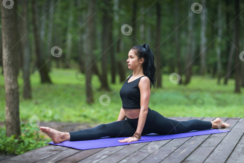 Скачать Молодая спортивная девушка, практикующая йогу, выполняющая упражнение хануманасана, продольный шпагат, тренируется, сидя на коврике на деревянной платформе в парке фотосток Ozero