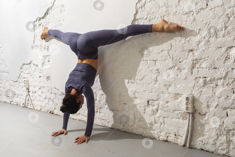Скачать Женщина, практикующая йогу, выполняет упражнение Адхо Мукха Врикшасана с Самаконасаной, стойкой на двух руках с поперечным шпагатом у кирпичной стены фотосток Ozero