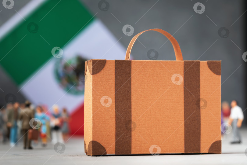 Скачать Картонный чемодан, пластмассовые игрушечные человечки и флаг на абстрактном фоне, концепция на тему переезда или миграции в Мексике фотосток Ozero