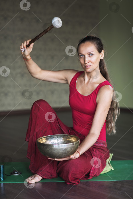 Скачать Женщина, практикующая йогу, выполняющая звуковую медитацию с металлической чашей, сидит на коврике в студии фотосток Ozero