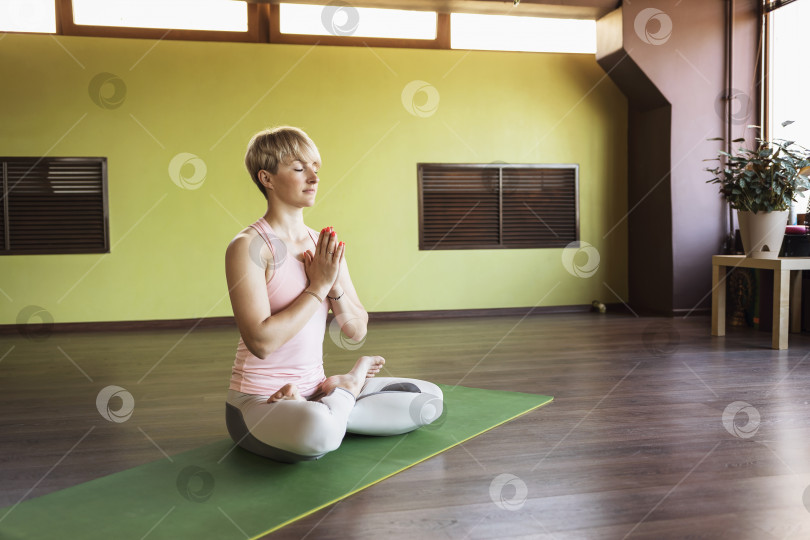 Скачать Женщина занимается медитацией в позе лотоса с намасте, сидя на коврике в студии фотосток Ozero