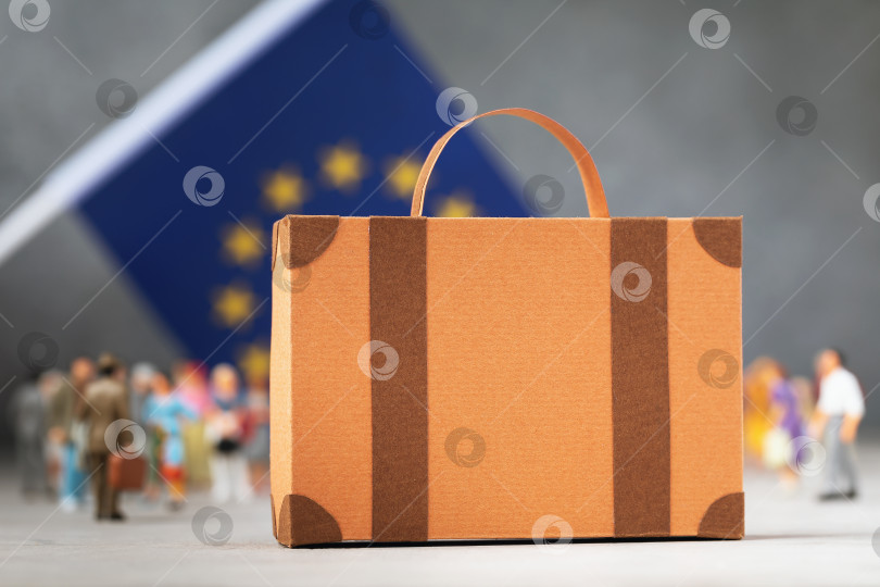 Скачать Картонный чемодан, пластмассовые игрушечные человечки и флаг на абстрактном фоне, концепция на тему переезда или миграции в Европейский союз фотосток Ozero