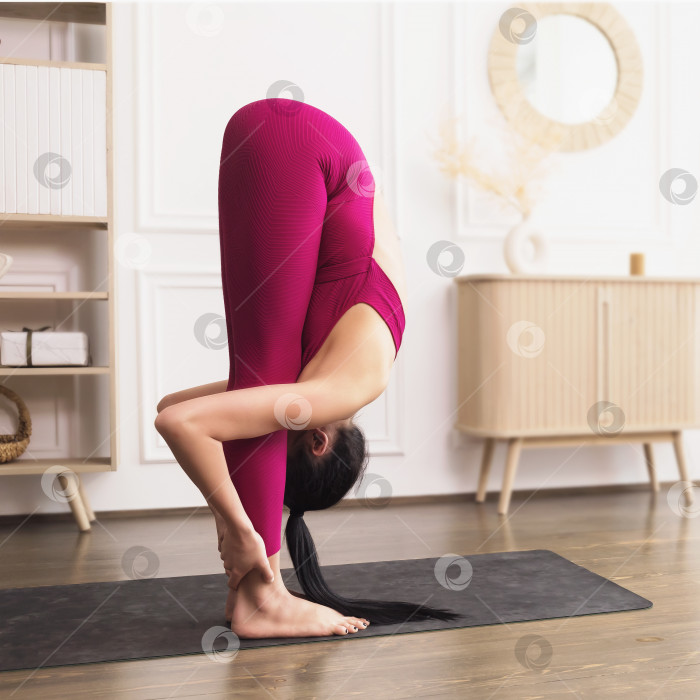 Скачать Спортивная молодая женщина, практикующая йогу, выполняющая упражнение уттанасана, наклоняясь вперед, касаясь головы ногами, тренируясь в спортивной одежде в комнате фотосток Ozero