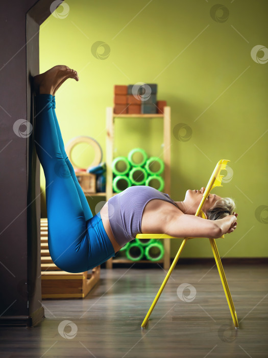 Скачать Женщина, ведущая здоровый образ жизни и практикующая йогу, выполняет наклон на спину на стуле, упираясь ногами в стену, тренируется в фитнес-центре в спортивной одежде, ракурс в полный рост фотосток Ozero