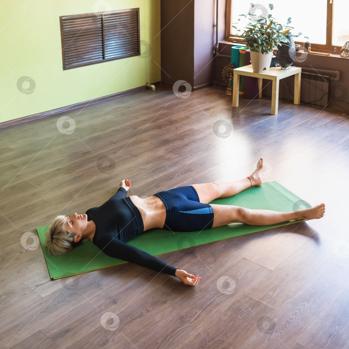 Скачать Женщина, практикующая йогу, выполняющая упражнение шавасана с дыхательной практикой пранаяма, расслабляющая асана, лежащая на коврике в комнате фотосток Ozero