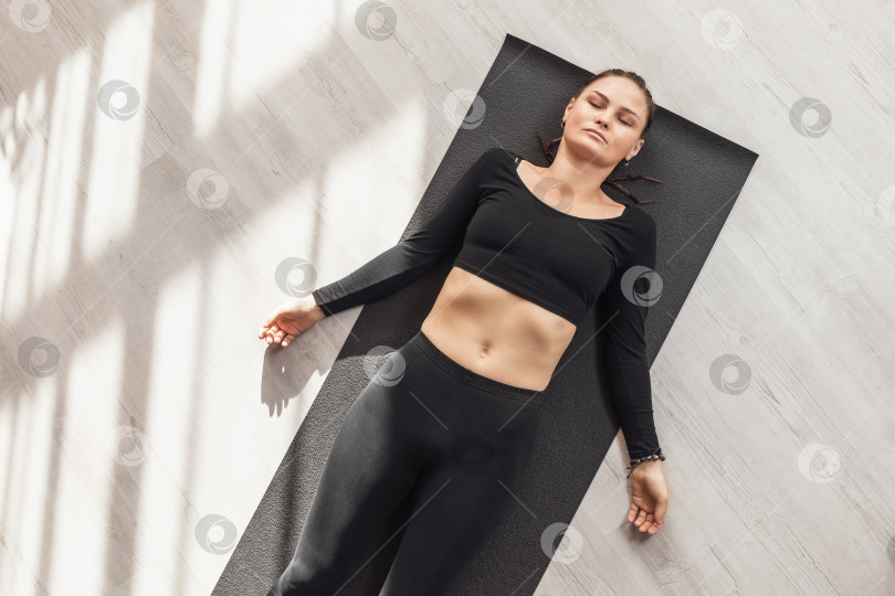 Скачать Женщина, практикующая йогу, выполняющая упражнение шавасана, поза расслабления, лежащая на коврике в спортивной одежде в комнате, вид сверху фотосток Ozero