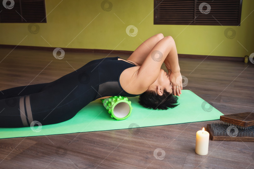 Скачать Женщина средних лет, практикующая йогу, выполняет расслабляющие упражнения для мышц спины с помощью массажного ролика, тренируется с закрытыми глазами, лежа на коврике в студии с намасте фотосток Ozero