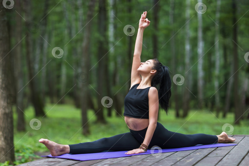 Скачать Молодая девушка, ведущая здоровый образ жизни и практикующая йогу, выполняет упражнение хануманасана, продольный шпагат, тренируется, сидя на коврике на деревянном мосту в парке фотосток Ozero