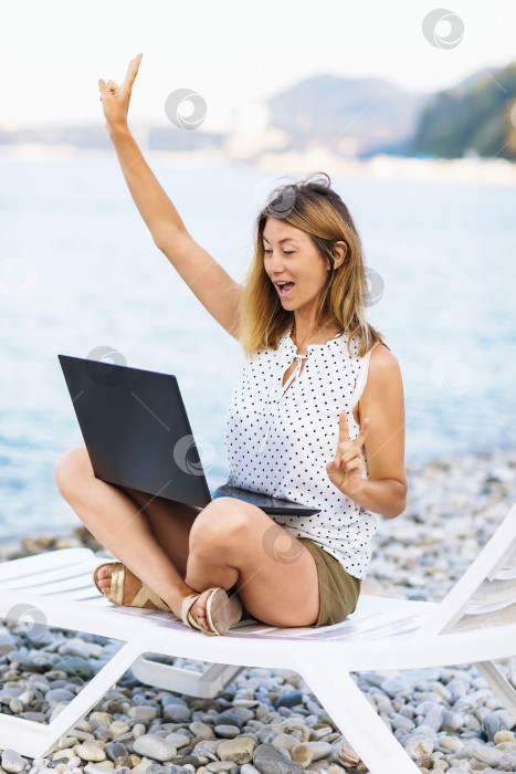 Скачать Женщина радуется заключению сделки, работая за ноутбуком, сидя в шезлонге на берегу моря, занимаясь онлайн-бизнесом, совмещая отдых и работу фотосток Ozero