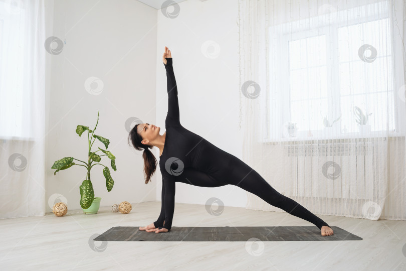 Скачать Женщина в черном цельном спортивном боди, практикующая йогу, выполняющая упражнение Уттхита Паршваконасана, поза с расширенным боковым углом, тренируется, стоя на коврике в комнате фотосток Ozero