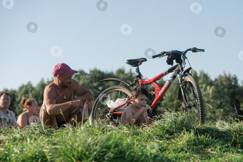 Скачать Россия, Гатчина, 20 июня 2022 года: Отец и его маленький сын отдыхают, сидя на траве с велосипедом фотосток Ozero
