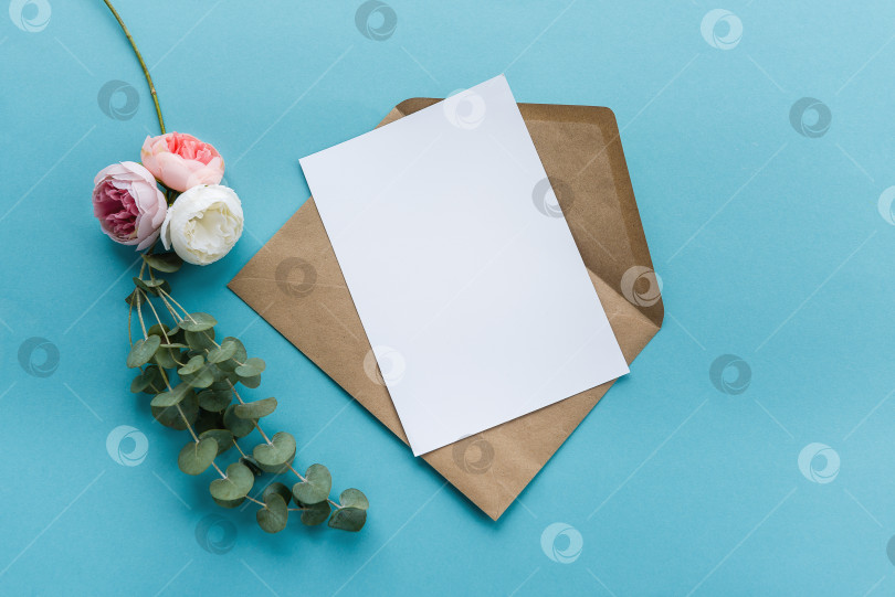 Скачать Конверт из крафт-бумаги с белой пустой карточкой, цветы на синем фоне. Подготовка открытки фотосток Ozero