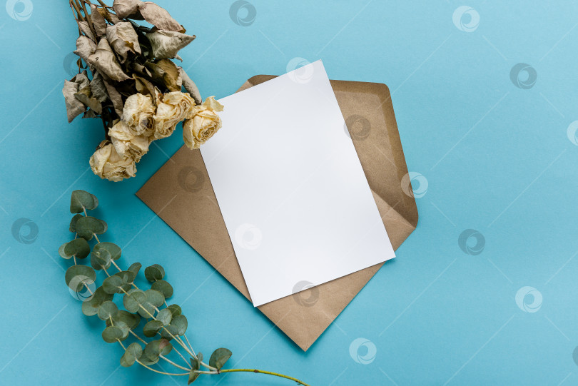 Скачать Конверт из крафт-бумаги с белой пустой карточкой, цветы на синем фоне. Подготовка открытки фотосток Ozero