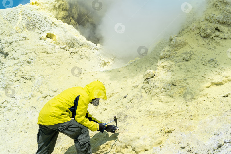 Скачать мужчина-вулканолог на склоне вулкана рядом с дымящейся серной фумаролой исследует образец минерала фотосток Ozero