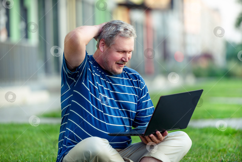 Скачать Счастливый мужчина просматривает онлайн-контент на ноутбуке и смеется, сидя на лужайке в городе фотосток Ozero
