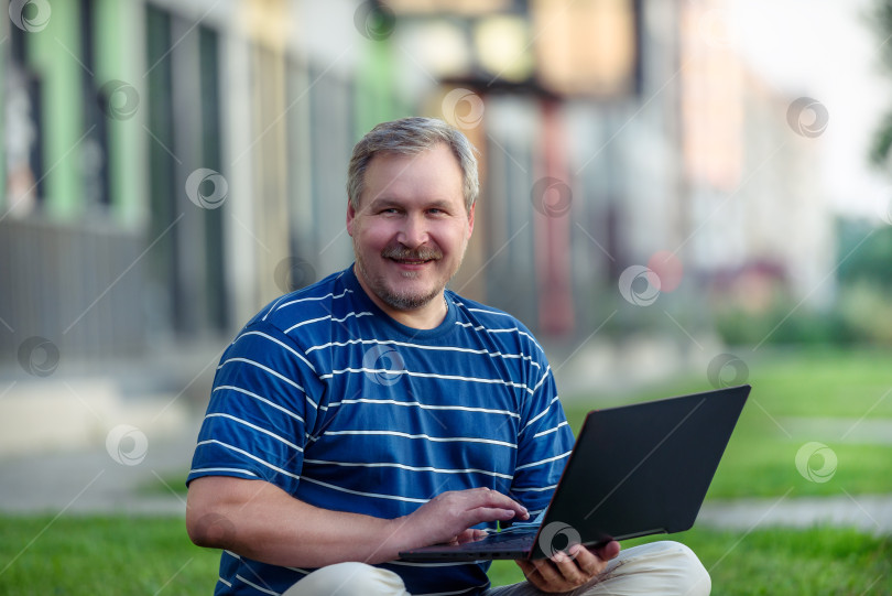 Скачать Счастливый человек, просматривающий онлайн-контент на ноутбуке, сидя на лужайке в городе фотосток Ozero