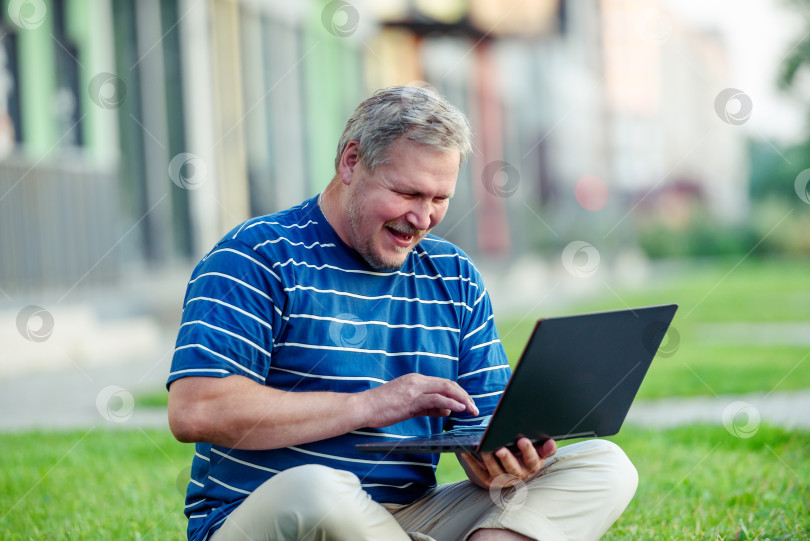 Скачать Счастливый мужчина просматривает онлайн-контент на ноутбуке и смеется, сидя на лужайке в городе фотосток Ozero