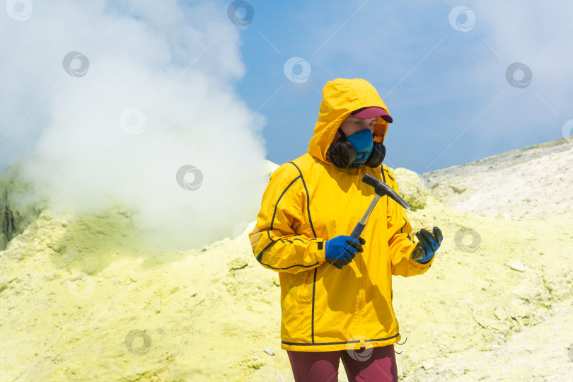 Скачать женщина-вулканолог на фоне дымящейся фумаролы исследует образец сернистого минерала геологическим молотком фотосток Ozero