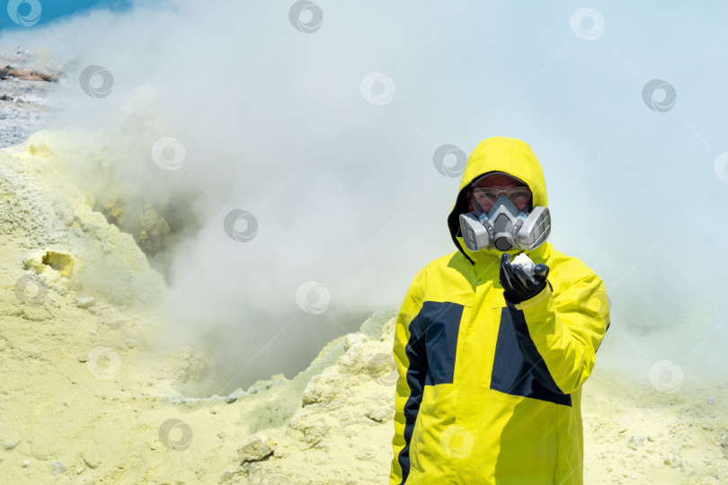 Скачать мужчина-вулканолог на фоне дымящейся фумаролы демонстрирует образец минерала фотосток Ozero