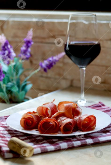 Скачать Тарелка с нарезанной прошутто и бокал красного вина на кухонном столе, фотография в стиле лайфстайл. Пшут и красное вино, итальянское или испанское вяленое мясо. Закуска из хамона с бокалом вина, концепция позднего завтрака фотосток Ozero