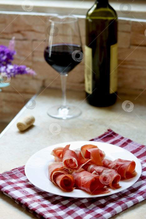 Скачать Тарелка с нарезанной прошутто и бокал красного вина на кухонном столе, фотография в стиле лайфстайл. Пшут и красное вино, итальянское или испанское вяленое мясо. Закуска из хамона с бокалом вина, концепция позднего завтрака фотосток Ozero