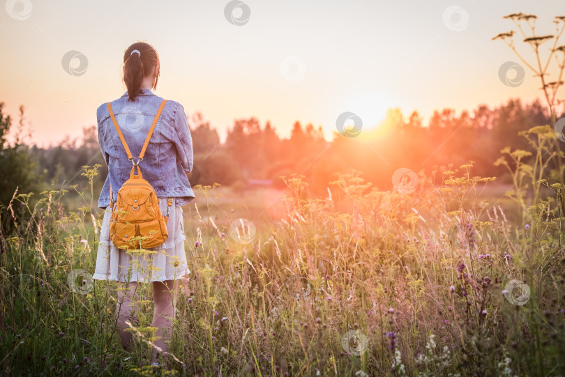 Скачать Молодая девушка в платье и с рюкзаком на природе на закате, вид сзади фотосток Ozero