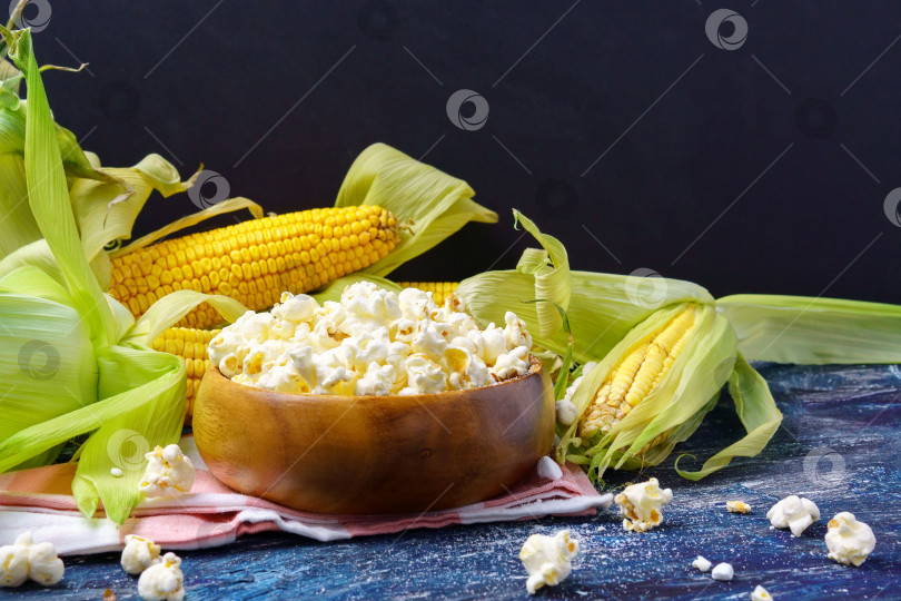 Скачать Вкусный попкорн в миске на столе. Початки свежей кукурузы на темном фоне. Копировальное пространство фотосток Ozero