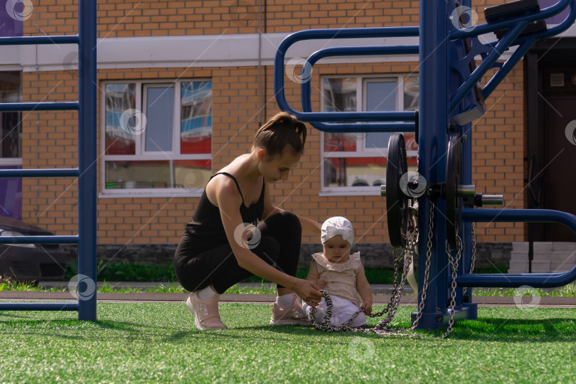 Скачать мама и малышка дочка гуляют на спортивной площадке возле дома, малышка интересуется тренажером фотосток Ozero