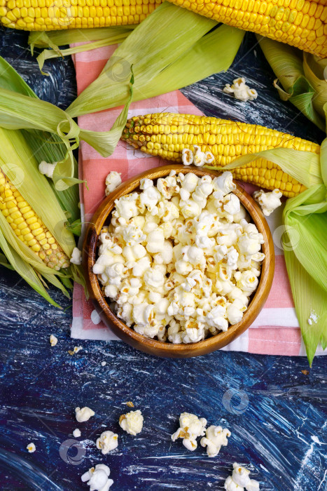 Скачать Вкусный попкорн в миске на столе. Початки свежей кукурузы на темном фоне. Вертикальная фотография фотосток Ozero