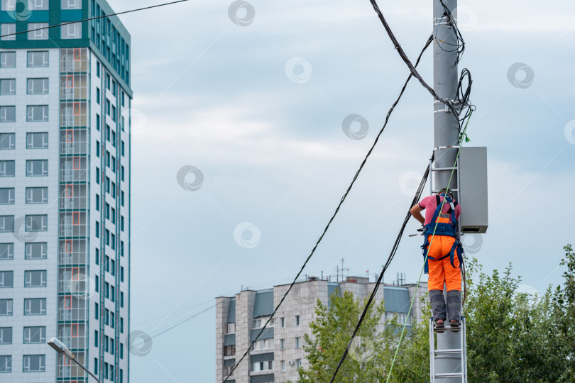 Скачать электрик или оператор линии связи работает над прокладкой кабеля на вершине телефонного столба на фоне городских зданий фотосток Ozero