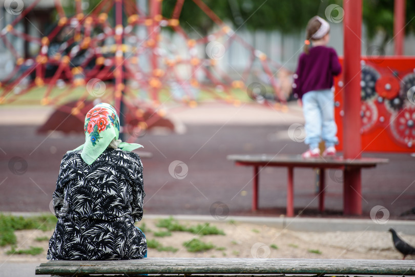 Скачать Россия, Гатчина, август 2020 года: бабушка сидит на скамейке и наблюдает, как ее внук играет на детской площадке фотосток Ozero