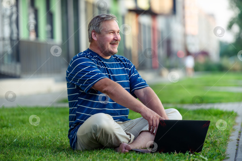Скачать Счастливый человек, просматривающий онлайн-контент на ноутбуке, сидя на лужайке летним днем в городе фотосток Ozero