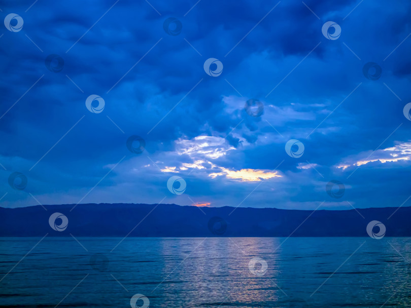 Скачать Облака над водой, освещенные заходящим солнцем. Погруженный в сумерки драматический пейзаж. фотосток Ozero