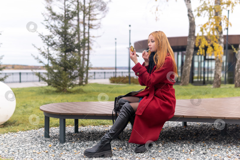 Скачать рыжеволосая молодая женщина поправляет свой макияж, сидя на скамейке в осеннем парке фотосток Ozero