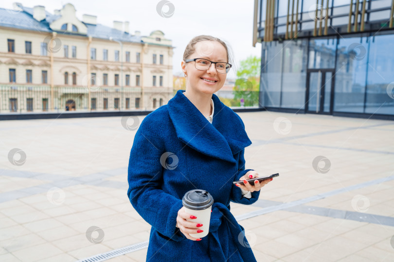 Скачать Деловая женщина с телефоном возле офиса. Портрет улыбающейся девушки в синем пальто фотосток Ozero