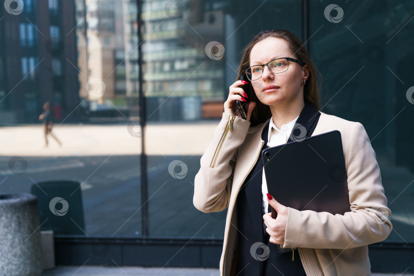 Скачать Деловая женщина в очках с ноутбуком в руках разговаривает по телефону возле бизнес-центра фотосток Ozero