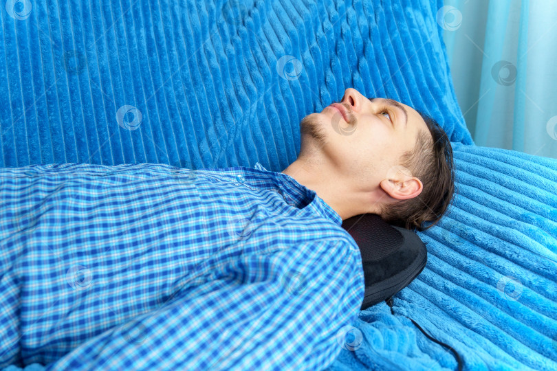 Скачать Мужчина лежит на диване и массирует себе шею электрической массажной подушкой. фотосток Ozero