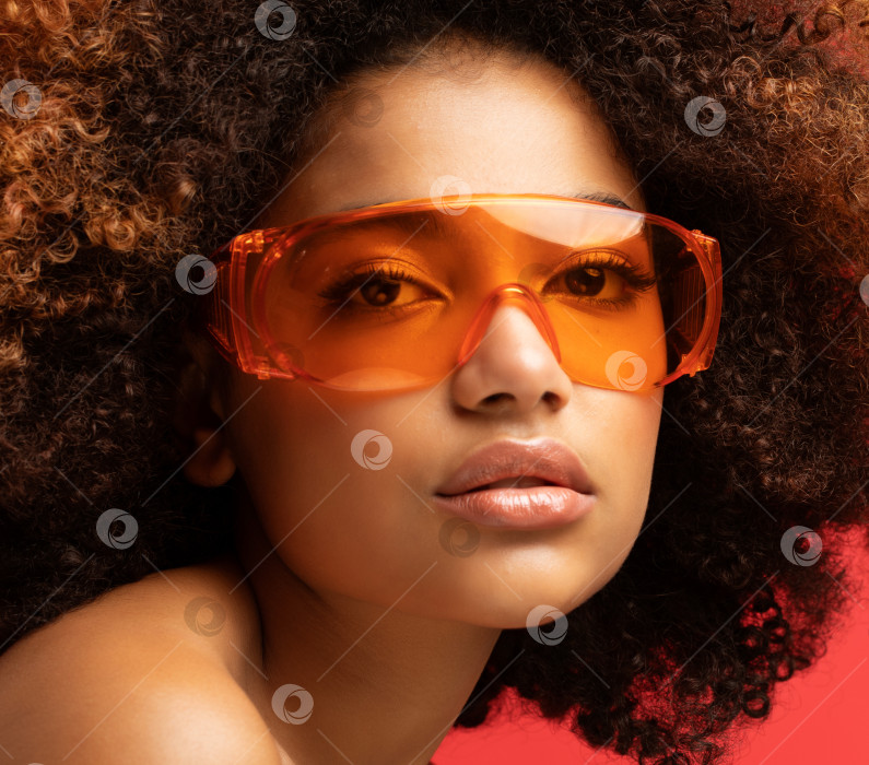 Скачать Молодая женщина в солнцезащитных очках, выделяющаяся на красном фоне студии. Возбужденная афроамериканка. фотосток Ozero