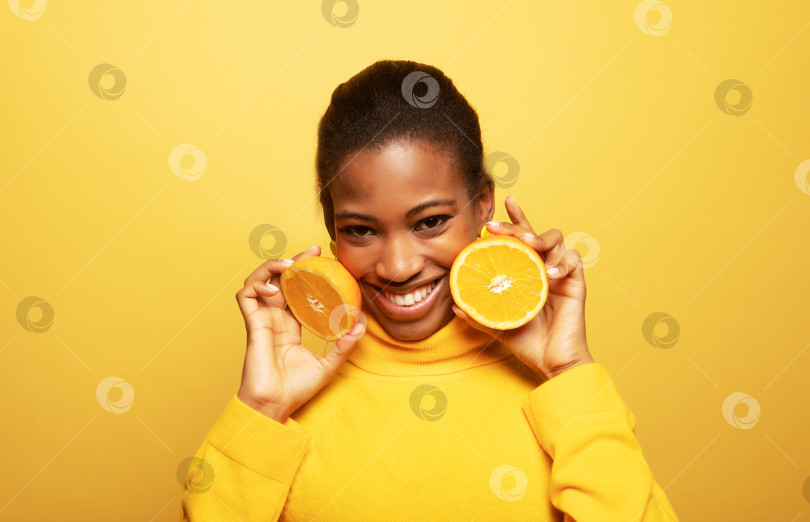 Скачать молодая афроамериканка, держащая в руках дольки апельсина, выглядит изумленно изолированной на фоне желтого цвета. фотосток Ozero