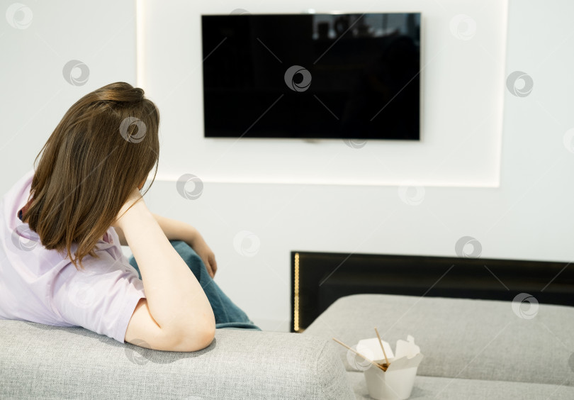 Скачать Женщина сидит на диване, смотрит телевизор, рядом стоит азиатская еда в коробке, одинокий вечер. фотосток Ozero