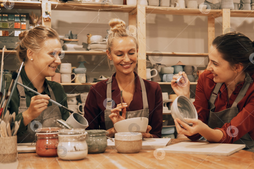 Скачать Компания из трех веселых молодых подруг занимается росписью керамики в гончарной мастерской. фотосток Ozero