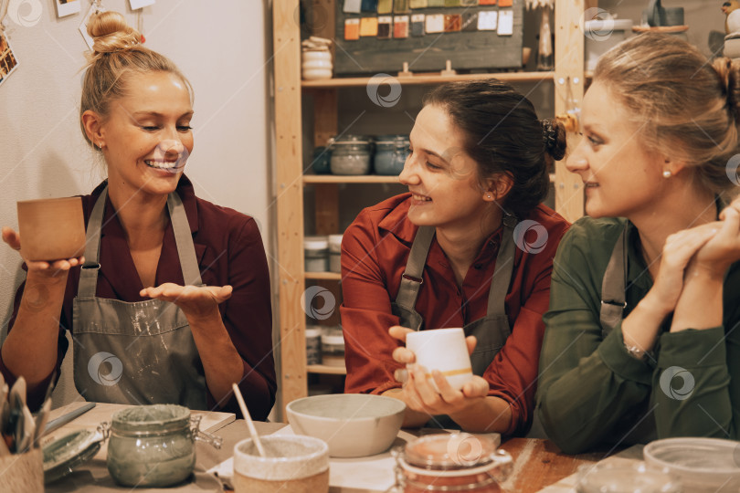 Скачать Компания из трех симпатичных молодых подруг изготавливает керамические кружки в гончарной мастерской. фотосток Ozero