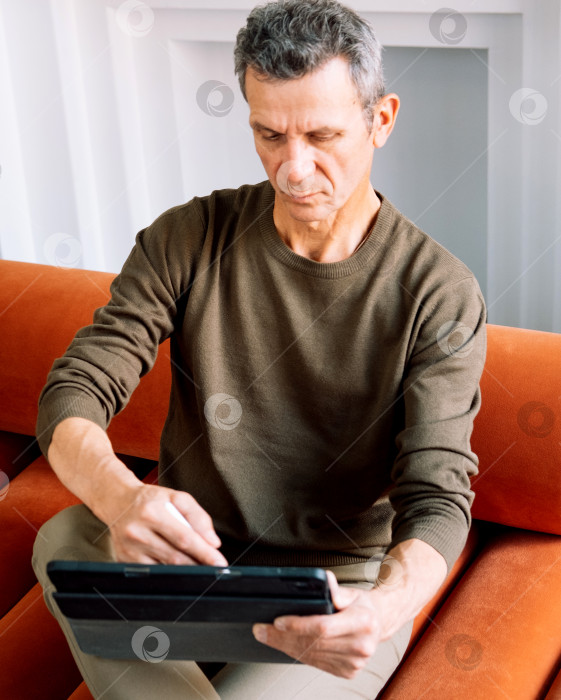 Скачать Жизнерадостный пожилой мужчина в повседневной одежде пользуется цифровым планшетом, сидя дома на диване фотосток Ozero