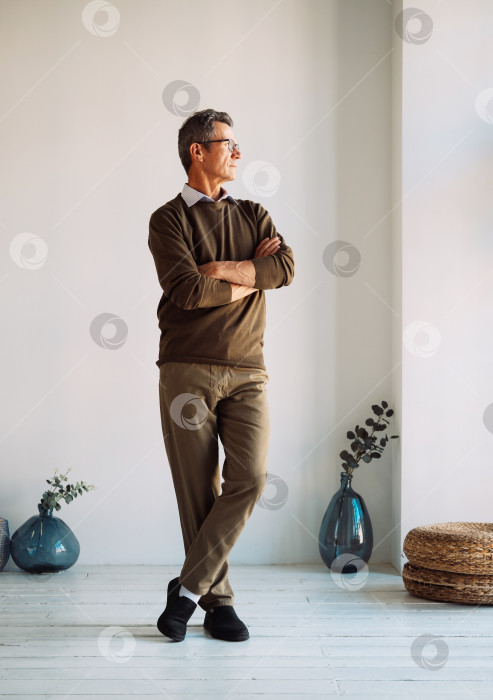 Скачать Уверенный в себе человек. Портрет жизнерадостного непринужденного пожилого мужчины, позирующего со скрещенными на груди руками, стоящего дома со скрещенными руками. фотосток Ozero