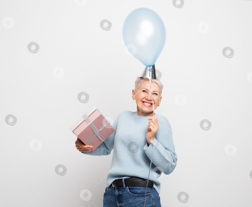 Скачать Удивленная пожилая женщина с короткими волосами в праздничной шляпе, держащая в руках голубой воздушный шар и подарочную коробку фотосток Ozero